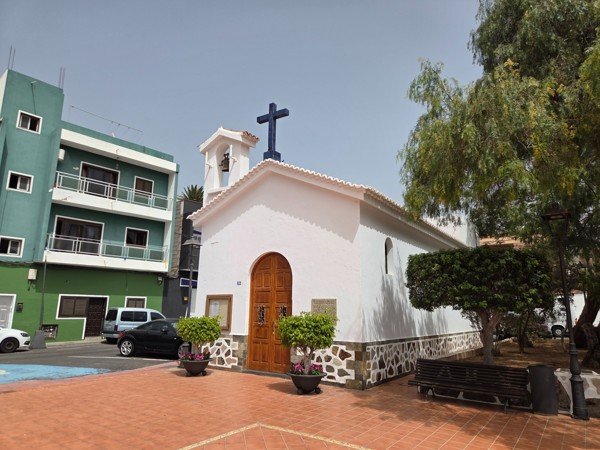 Small white chapel of Aldea Blanca with a bell gable, wooden door and shaded benches.