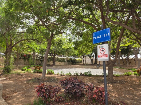 Blue street sign for Plaza-Era marking a shaded square with trees and low borders in Aldea Blanca.