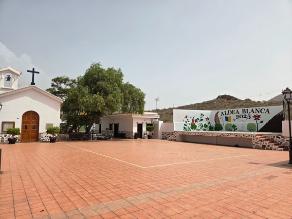Open tiled square beside the white village church of Aldea Blanca with a stage mural and low hills behind.