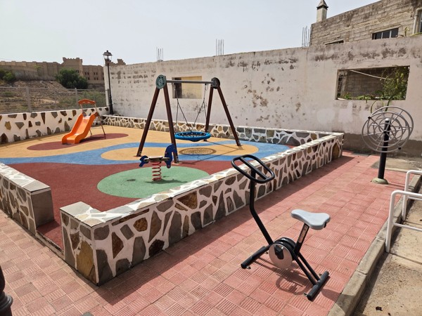 Compact children’s playground in Aldea Blanca with slide, spring toy and circular swing; castle walls visible behind.
