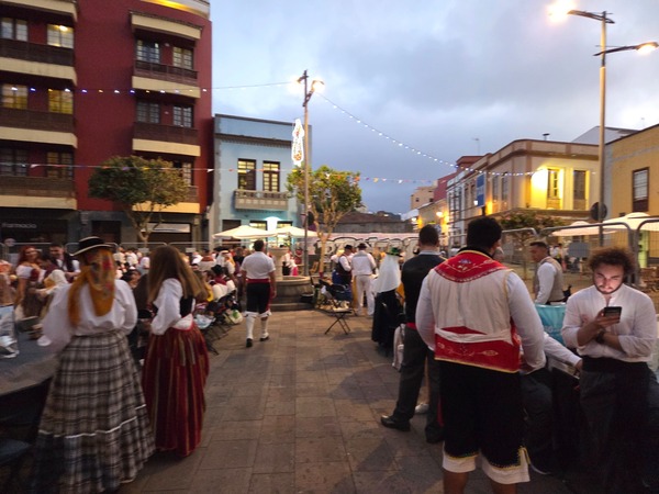 Festive crowd at dusk under strings of lights, many wearing traditional outfits, chatting around long tables.