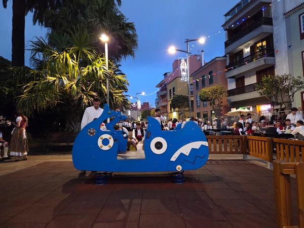 Children's play area with a blue rocker while people in traditional clothing sit and talk in the background.
