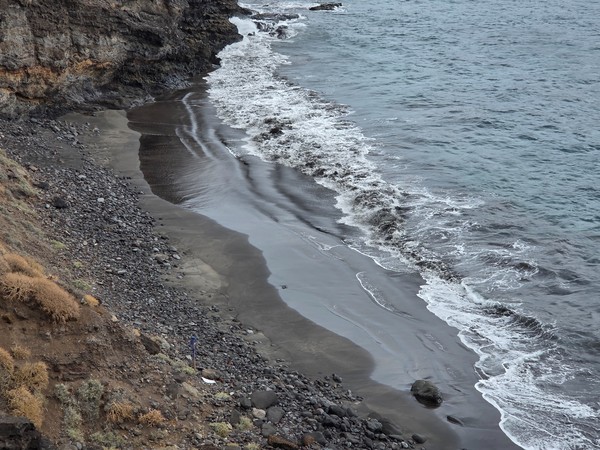 Remote black sand beach at the mouth of Barranco de Zapata with waves washing the shoreline below steep volcanic cliffs.