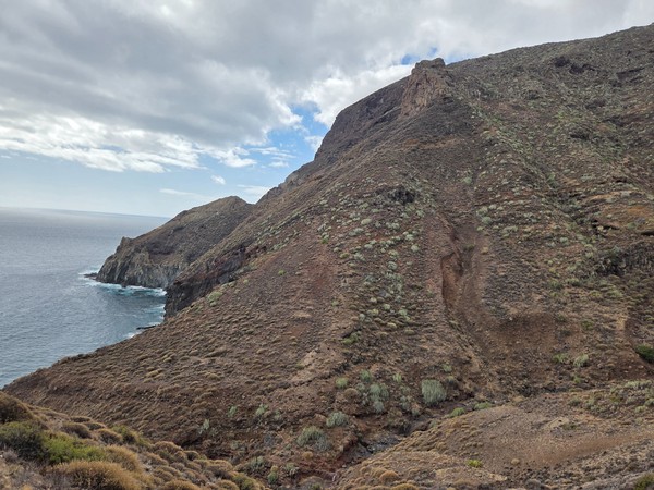 Wide view of rounded mountains around Barranco de Zapata with sparse green vegetation and the Atlantic ocean in the background.