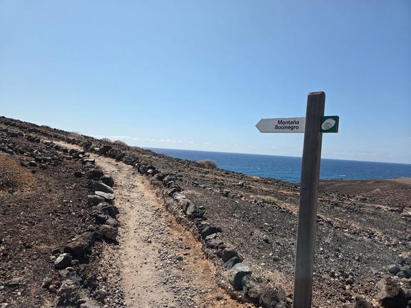 Trail sign pointing toward Montaña Bocinegro with the ocean in the background and a volcanic path.