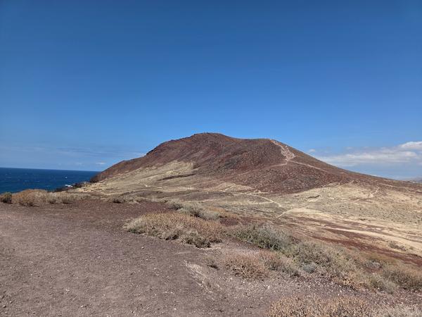Trail winding up Montaña Roja, visible with volcanic red and beige layers under a clear blue sky.
