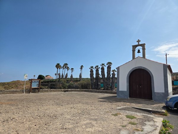 Small white chapel with arched door and bell tower in rural surroundings.
