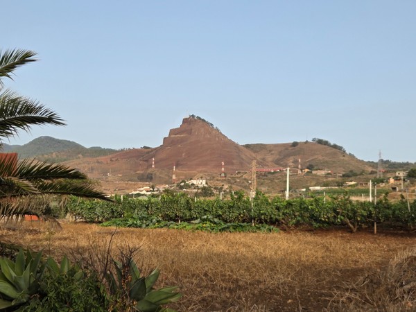 Distinctive red volcanic hill with stepped terraces and antennas, rising above farmland near the Camino Viejo de Candelaria.