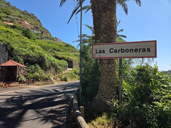 Entrance road sign to Las Carboneras village, framed by palm trees and mountain greenery.