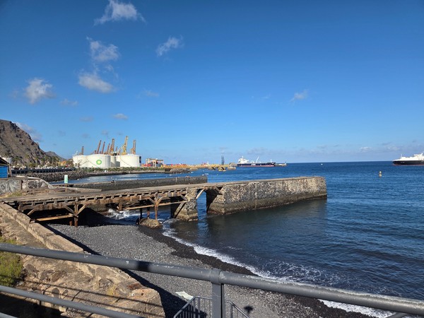 Panoramic view from Los Charcos toward the port with storage tanks, cranes and a calm pebble beach.