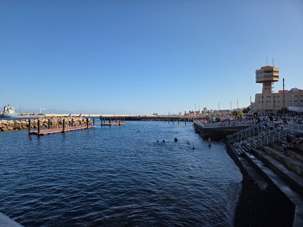 Wide overview of Los Charcos de Valleseco showing the floating docks inside the breakwater and swimmers enjoying calm water.