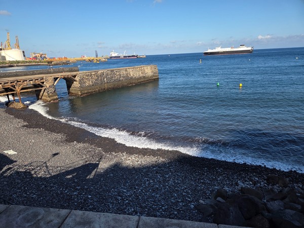 Rocky shoreline of Los Charcos with the old wooden pier and a ferry offshore beyond the breakwater.