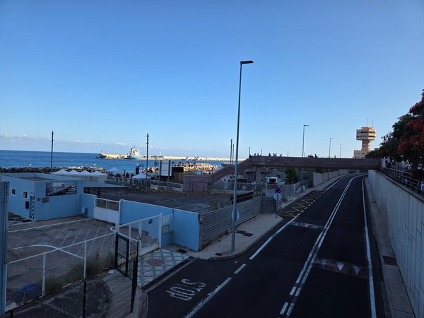Two-lane access road leading to Los Charcos with a pedestrian footbridge and the harbour control tower in the distance.