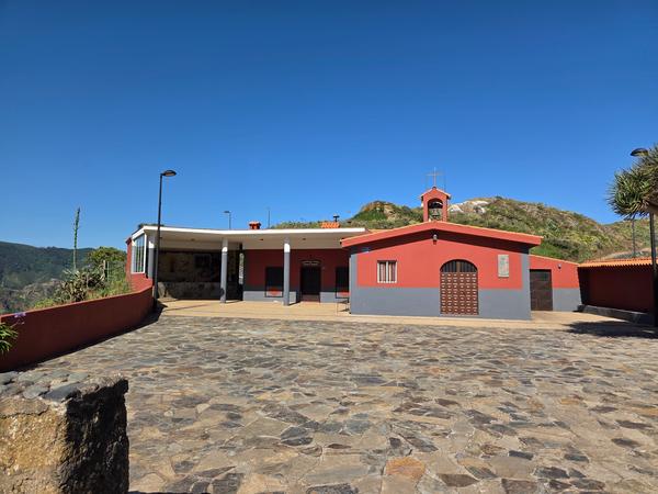 Small red and grey church with a bell tower at Chinamada village plaza, surrounded by stone-paved ground.