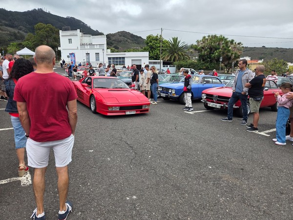 Red Ferrari sports car arriving slowly between parked classics as spectators gather around during the Tegueste show.