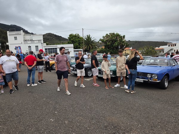 Line of classic Alfa Romeo coupes in blue, green and dark grey, parked nose-to-nose while visitors chat in front of them.