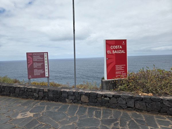 Entrance boards for Costa El Sauzal showing safety icons and opening hours beside a low lava-stone wall.