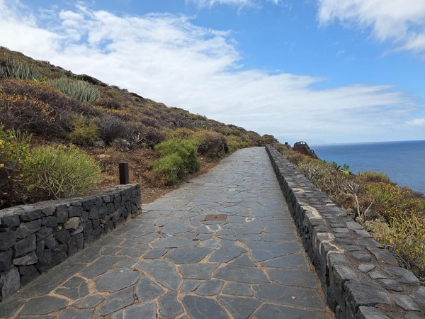 Broad paved promenade bordered by volcanic stone walls and coastal vegetation.