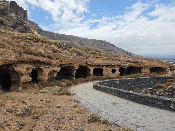 Row of shallow sea caves in honeycombed tuff beside the path, with the cliff rising above.