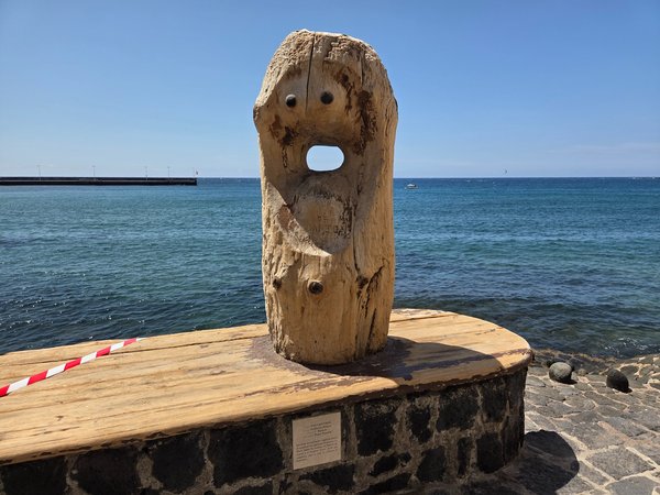 Whimsical driftwood figure called ‘Encuentros’ standing on a stone plinth by the water.