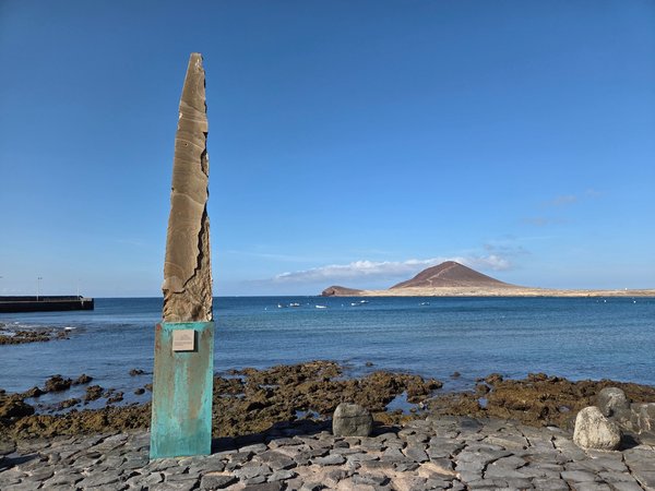 Wide coastal view towards Montaña Roja with a tall stone sculpture in the foreground at El Médano's seafront.