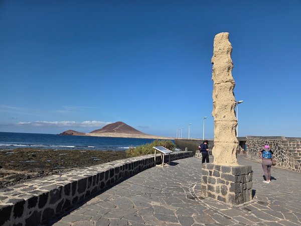 Textured vertical column sculpture framing Montaña Roja in the distance along the seaside path.