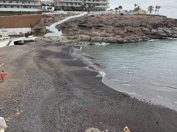 Black-sand shoreline of Playa El Pinque with pebbles and gentle waves at low tide.