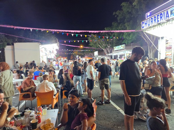 People dining at outdoor tables near a churrería and brightly lit food stalls during the Fiestas de El Fraile 2025.
