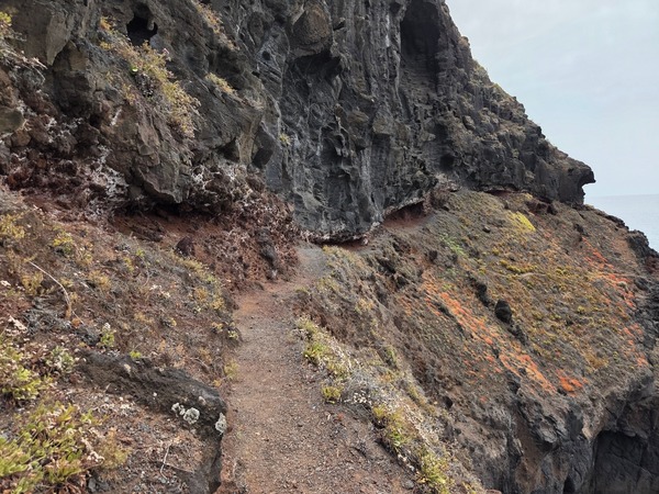 Narrow hiking path carved into the cliff on the descent to Playa de Garañona, with rough lava overhangs above.