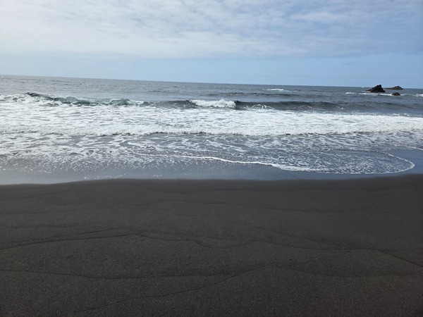 Small set of Atlantic waves washing across the black sand at Garañona during calm conditions.