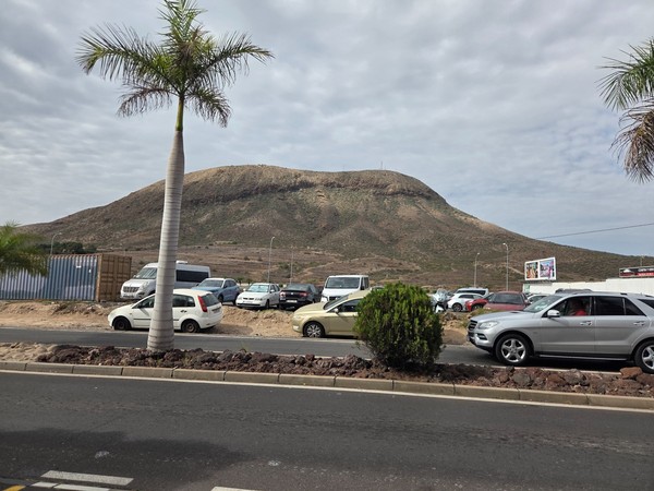 View of the volcanic Montaña de Guaza in Tenerife rising behind a parking area with palm trees and cars.