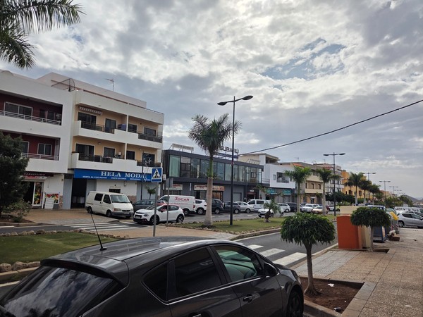 View of the main street of Guaza in Tenerife with apartments, shops, cars and palm trees under a cloudy sky.