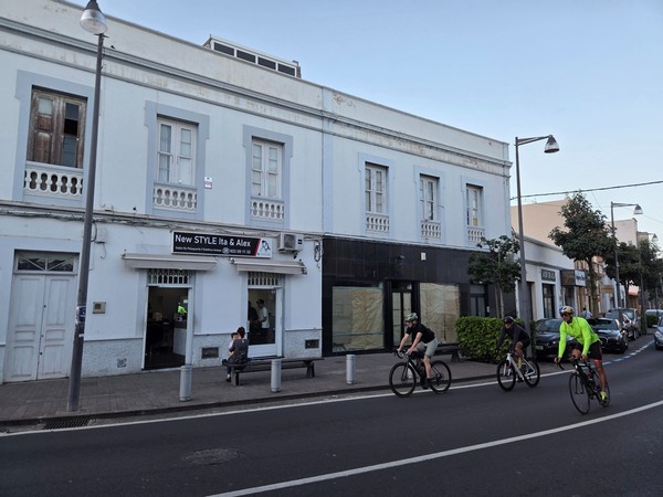 Cyclists riding along a central street in Güímar past a hair salon and people sitting on a bench.