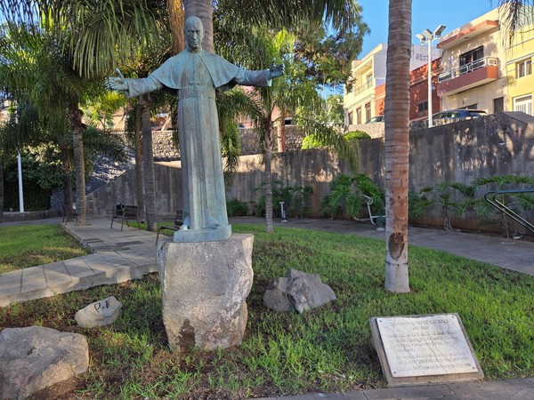 Bronze statue of a priest with open arms in a small park in Güímar surrounded by palm trees and benches.