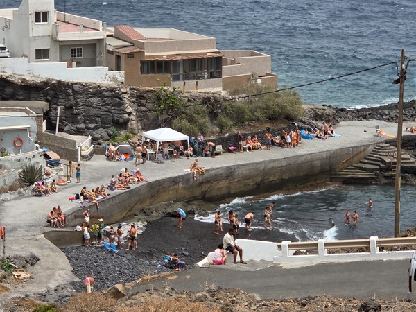 Families relax on the concrete platform and black pebble beach of La Caleta de Punta Prieta while children play in the shallow cove.