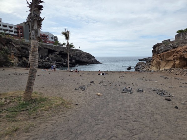 Quiet black-sand beach at Playa Las Galgas with scattered pebbles and low waves between rocky headlands.