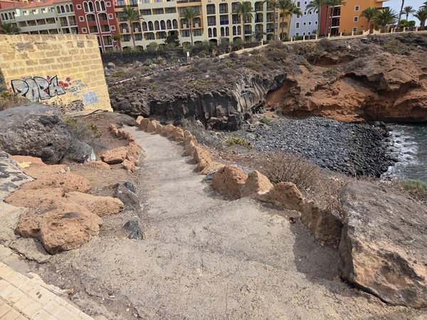 Narrow concrete path with lava-stone edges descending from the promenade toward Playa Las Salinas.