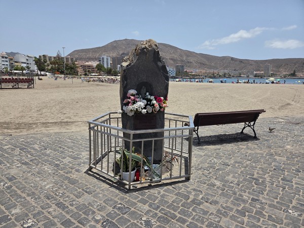 Stone memorial with flowers and candles standing on the promenade of Playa de Los Cristianos facing the beach.