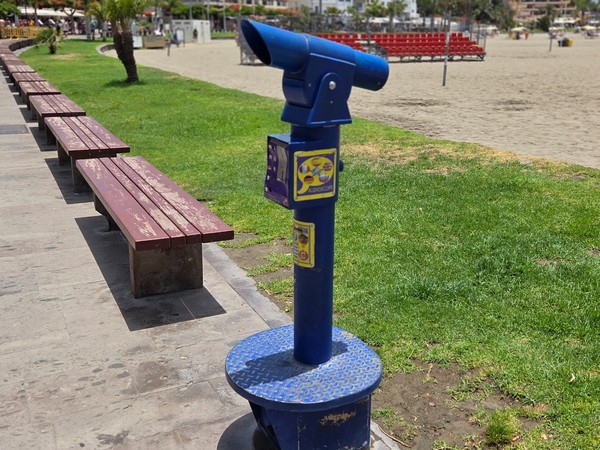 Blue telescopic viewer installed beside benches and green grass along the promenade of Playa de Los Cristianos.