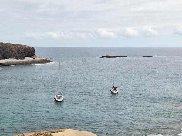 Two sailboats anchored near Playa de los Morteros with a rocky coastline in the background.