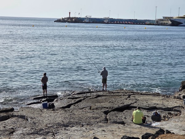 Two anglers standing on the rocks at Playa de Los Tarajales with fishing rods pointed towards the calm sea and the Los Cristianos harbour and ferry in the background.