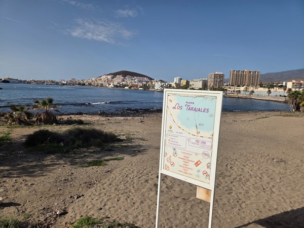 Information sign for Playa de Los Tarajales on the sand, with a map of the beach, safety icons and the town of Los Cristianos and Montaña de Guaza in the background.