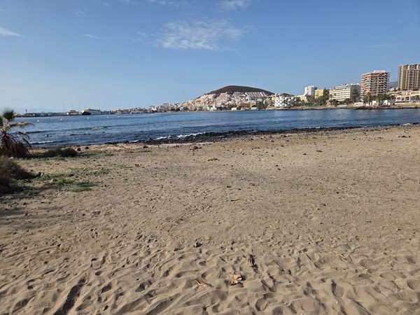 Open stretch of dark sandy beach at Playa de Los Tarajales with footprints in the sand and the town of Los Cristianos and harbour ferries visible across the bay.