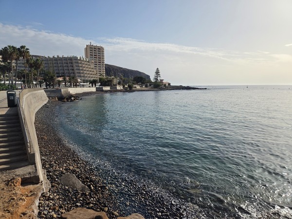 Wide panoramic view of Playa de Los Tarajales with the sandy foreground, calm sea and Montaña de Guaza rising behind the buildings of Los Cristianos in Tenerife.