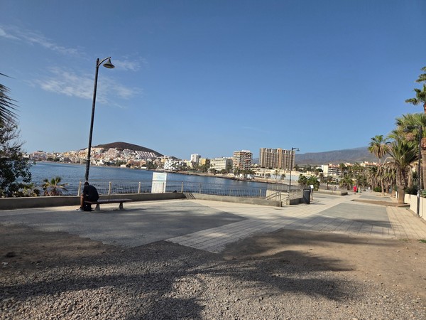 Promenade at Playa de Los Tarajales with a lone person sitting on a bench and Montaña de Guaza and the buildings of Los Cristianos across the bay.