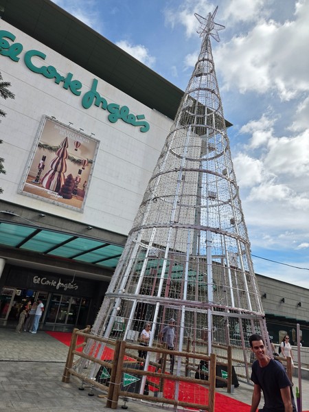 Large metallic Christmas tree in front of the El Corte Inglés building in Tenerife with visitors walking along the red carpet.
