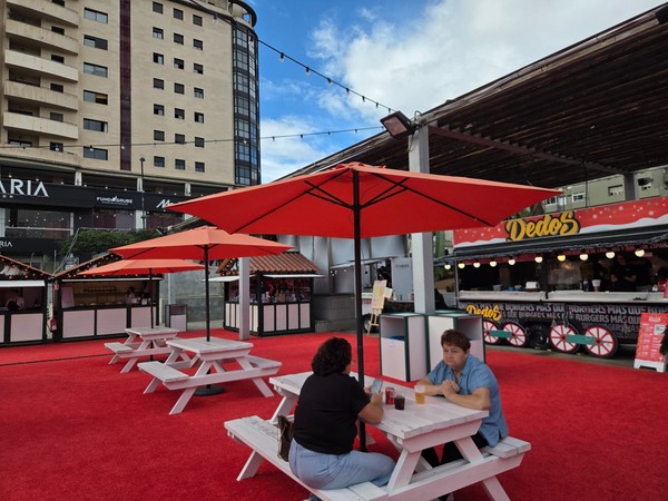 Outdoor dining area with white picnic tables, orange umbrellas and a food stand at the El Corte Inglés Christmas market in Tenerife.