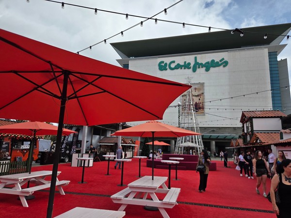 Wide view of the Christmas market in front of El Corte Inglés Tenerife with red carpet, umbrellas, picnic tables and the tall metallic tree.