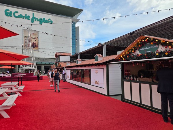 Line of decorated Christmas stalls in front of El Corte Inglés with people walking along the red carpet at the outdoor market in Tenerife.