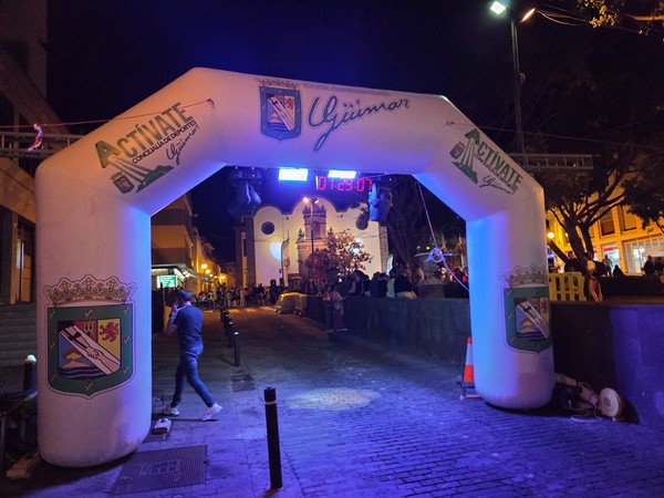 Inflatable race arch of La Nocturna Güímarera lit up at night on the main street of Güímar with people waiting around.